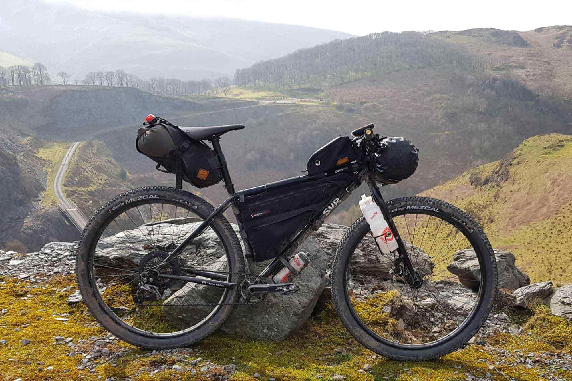 Mountain bike loaded with bags, frame pack, and multiple water bottles, leaning against rock on mountain overlook