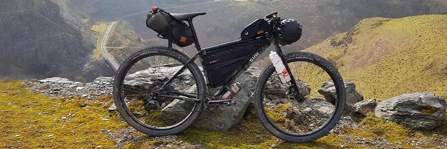 Mountain bike loaded with bags, frame pack, and multiple water bottles, leaning against rock on mountain overlook