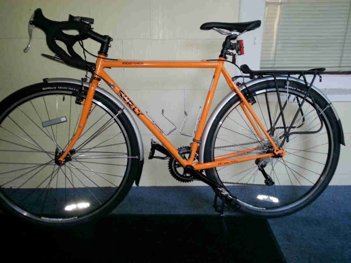 Left side view of an orange Surly Cross Check bike, parked on a black mat and blue carpet, in front of a white wall