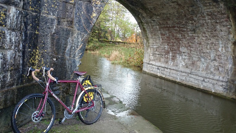 Left side view of a purple Surly Disc Trucker bike, parked on a stone wall, in front of a brick tunnel over a stream