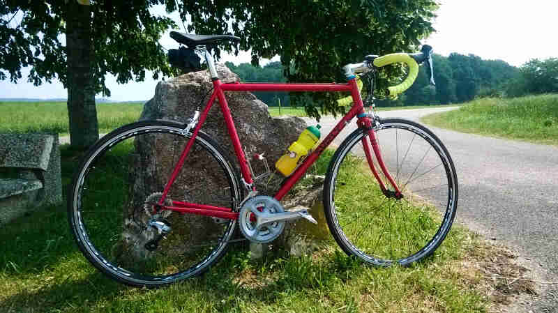 Right side view of a red Surly Pacer bike, parked in grass, against a rock, next to a paved trail