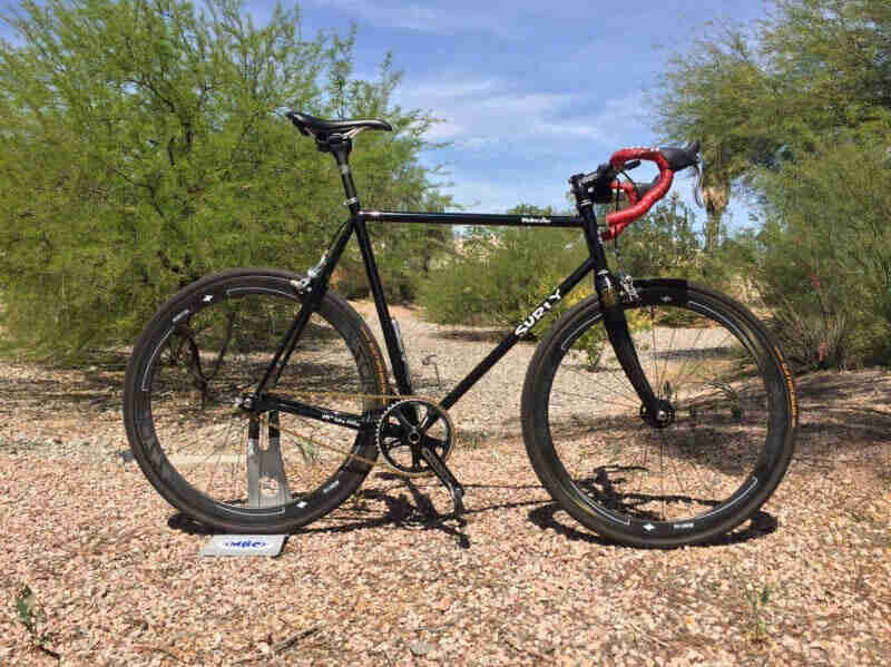 Right profile of a Surly Steamroller bike, black, parked in red desert gravel, with green bushes in the background