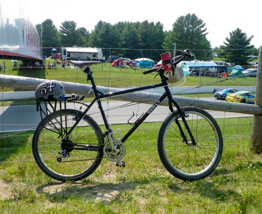 Right side view of a black Surly Troll bike with a helmet on the rear rack, leaning a wire fence at a racecar track