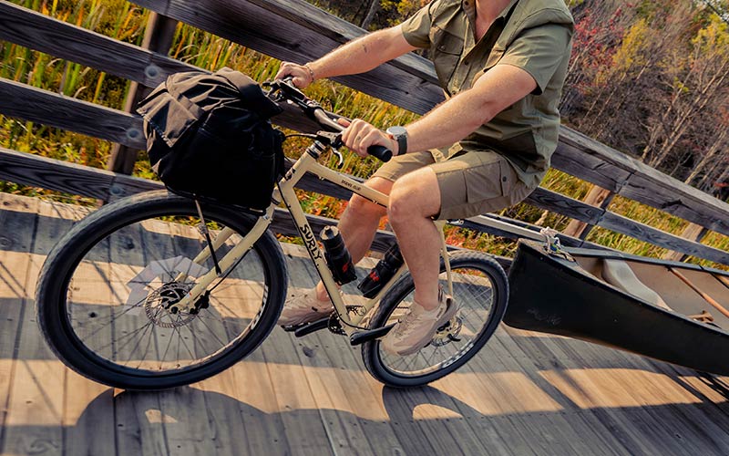 Cyclist riding Surly Bridge Club across wooden bridge towing canoe focus on bike