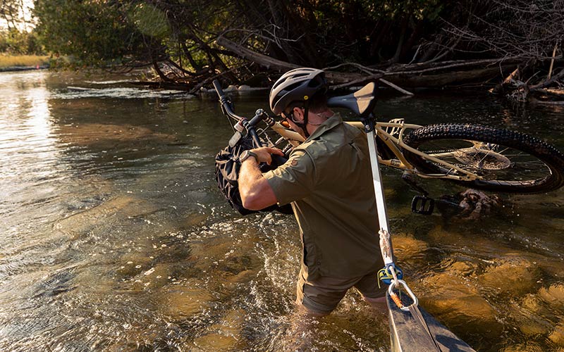 Cyclist shouldering Surly Bridge Club bike wih front rack bag with canoe in tow walking through water