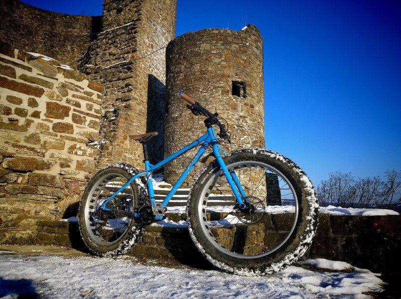 Right side view of a blue Surly fat bike, parked on snow, against the stone steps of a crumbling castle