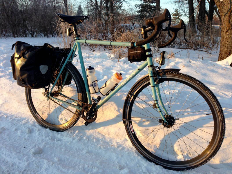 Right side view of a mint Surly Cross Check bike with rear saddlebags, parked along a snowy trail, with woods behind