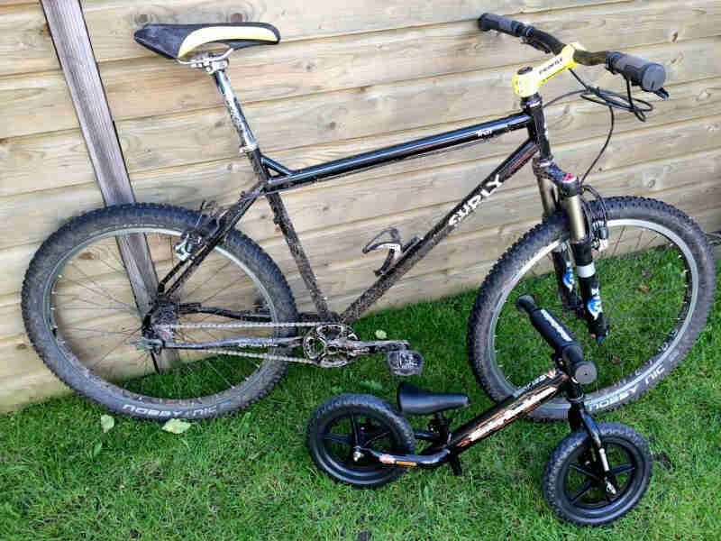 Right side view of a black Surly Troll bike, parked on grass, in front of a wooden wall with a child's bike below