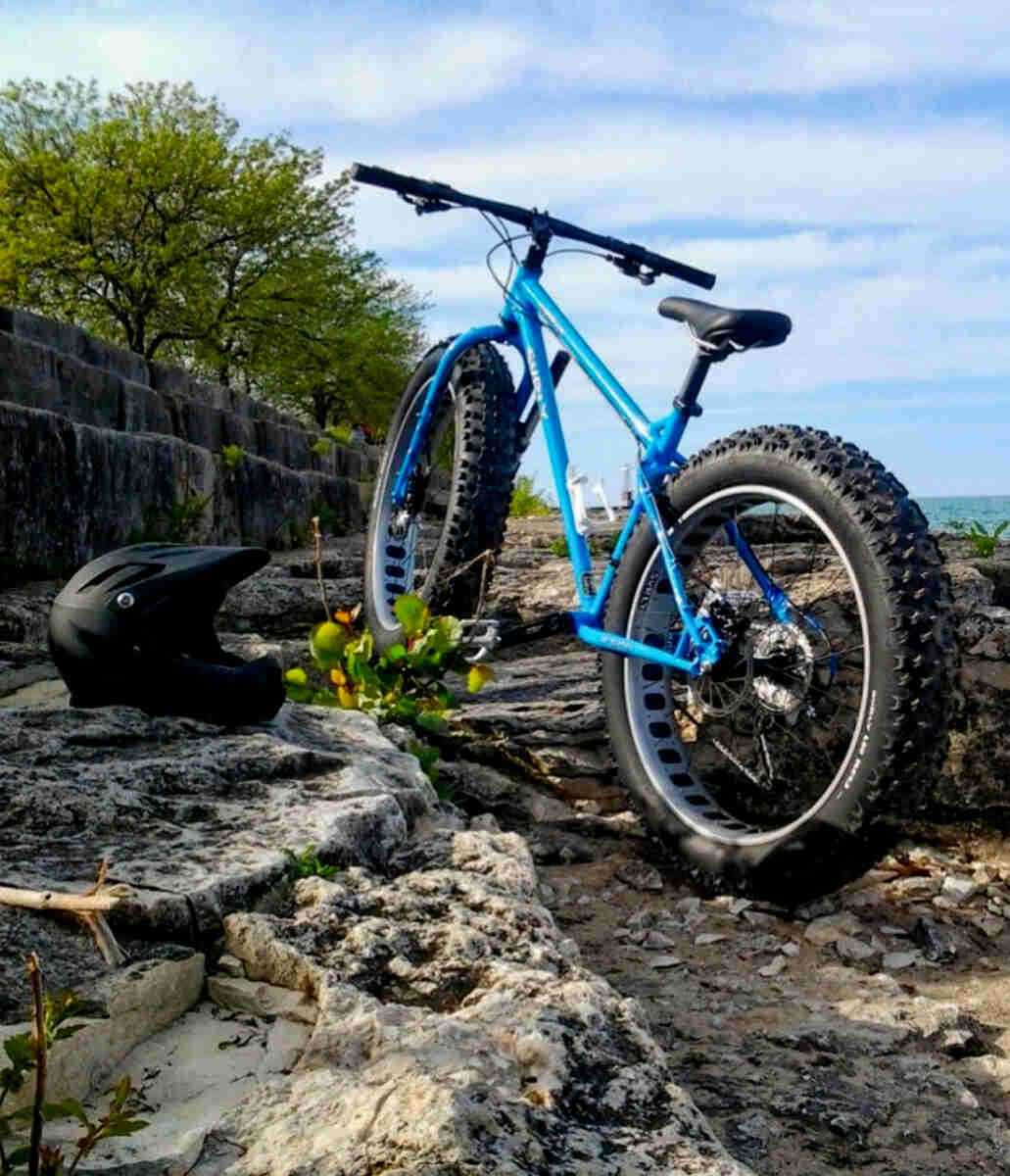 Rear, left side view of a blue Surly Ice Cream Truck fat bike, parked on a flat rock formation, next to a black helmet