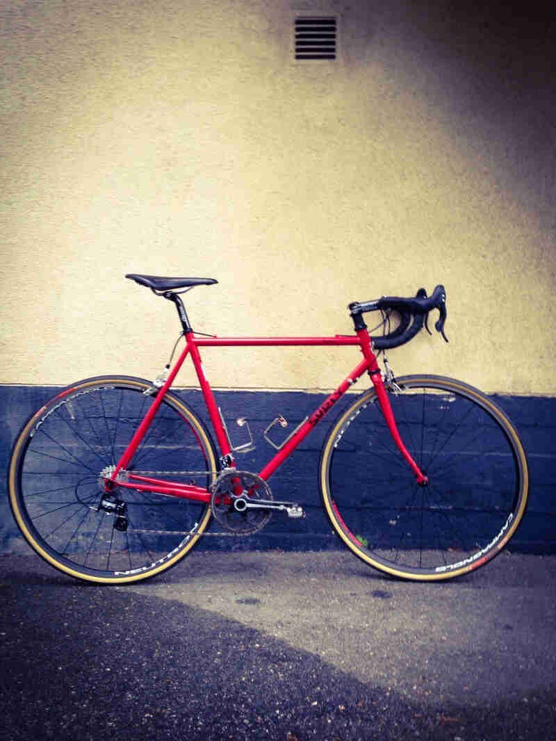 Right side view of a red Surly Pacer bike, parked on blacktop, leaning against a stucco and brick wall