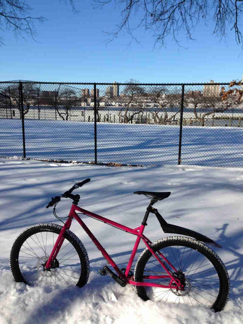 Left side view of a pink Surly bike, standing in deep snow, next to a chain link fence with a playing field behind it