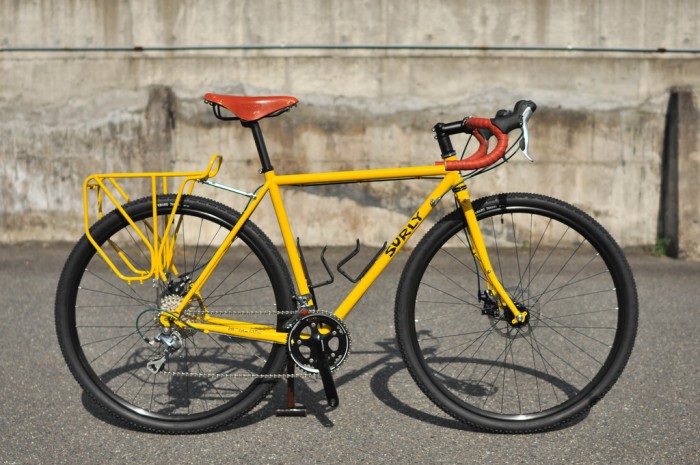 Right side view of a yellow Surly Straggler bike with rear rack, parked on pavement with a cement wall in the background