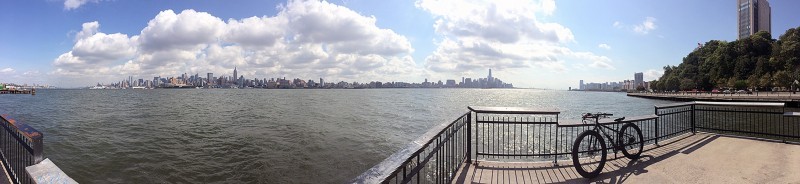 Panoramic, left side view of a Surly bike, against the handrail of a concrete dock, with a bay and city skyline behind