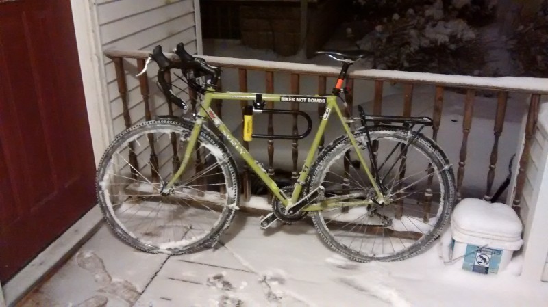 Left side view of a lime green Surly bike, parked on the handrail on a snowy porch, next to a house door at nighttime