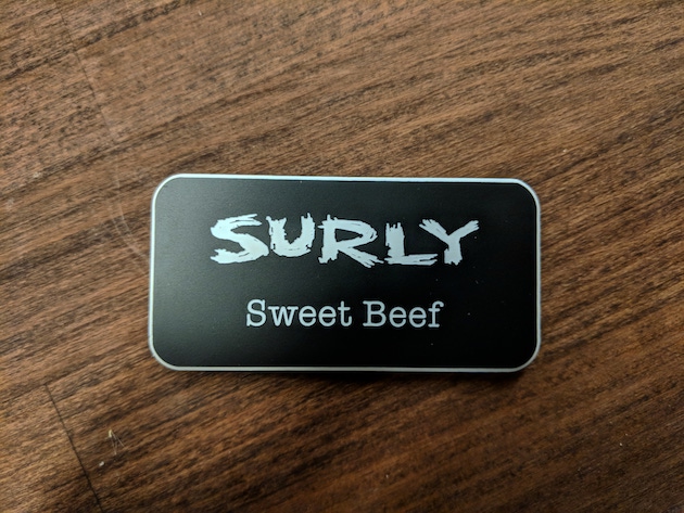 Black Surly name tag with Sweet Beef in white lettering lays on a wood surface