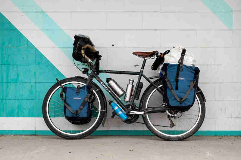 Left side view of a black Surly Disc Trucker bike with gear, parked on a walkway, against a white and teal wall