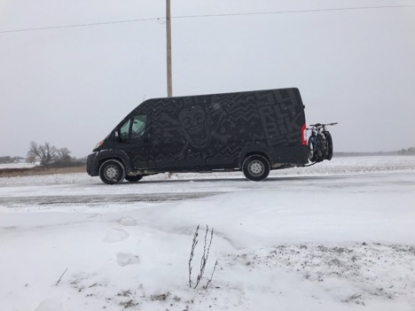 Left side view of a black cargo van with bikes on the back, driving on a snowy road