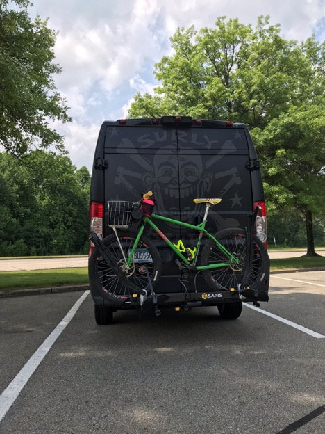 Right side view of a green Surly bike, mounted on the rear of a black van, in a parking lot with trees behind