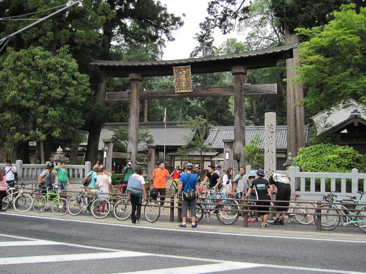 People and bikes lined up along a railing, in front of a torii arch with trees and pagoda buildings behind it