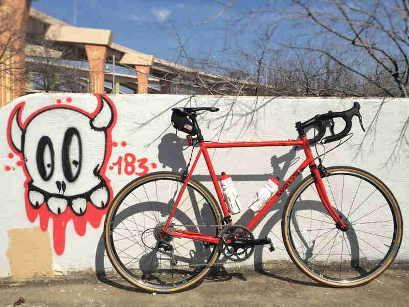 Right side view of a red Surly bike leaning on a cement wall with graffiti painted on it