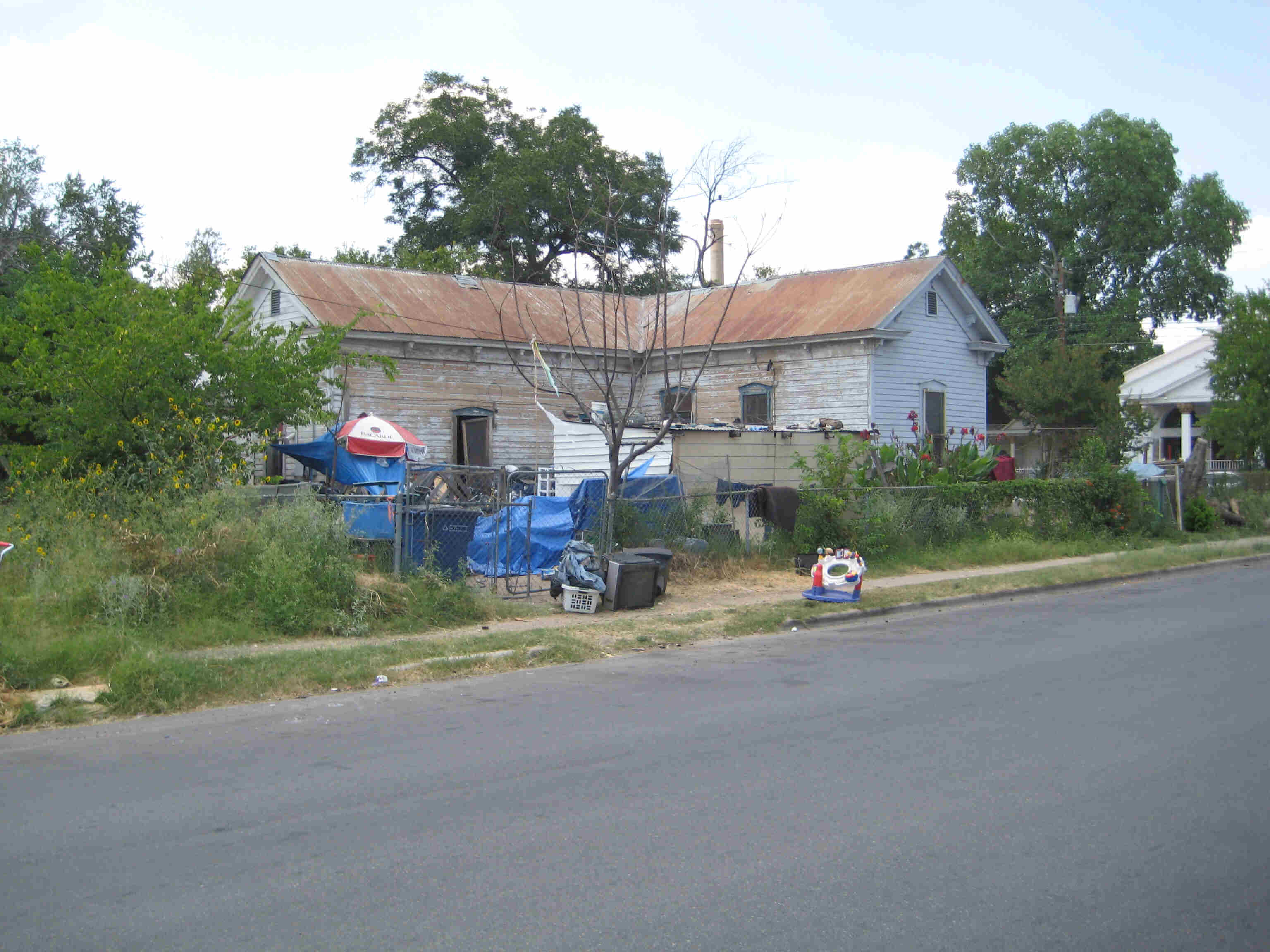 Street view of a house with a rusty steel roof, with a fenced yard full of weeds and junk