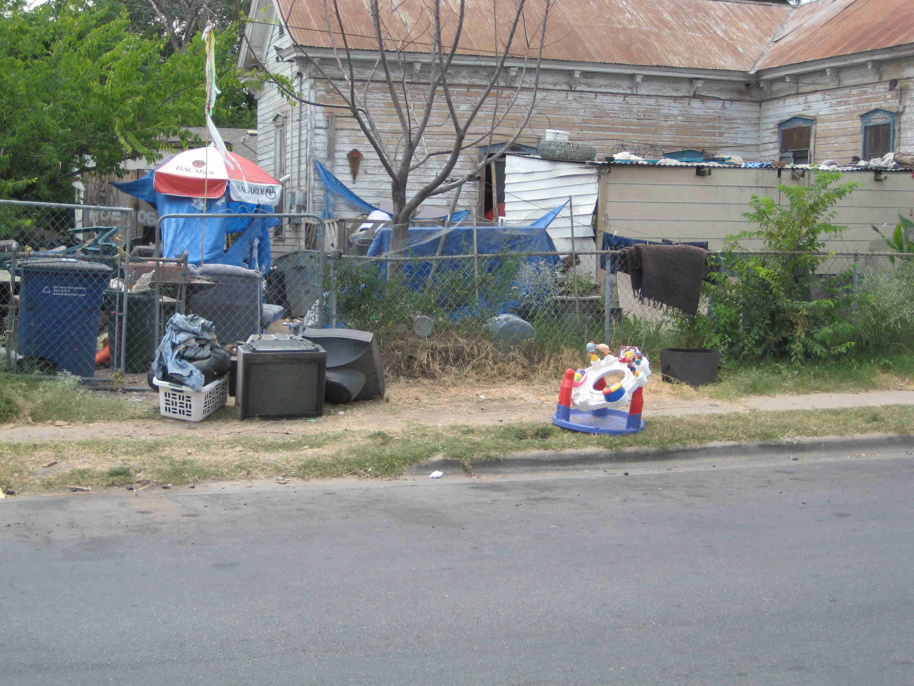 Side, street view of a house with a rusty steel roof, with a fenced yard full of weeds and junk