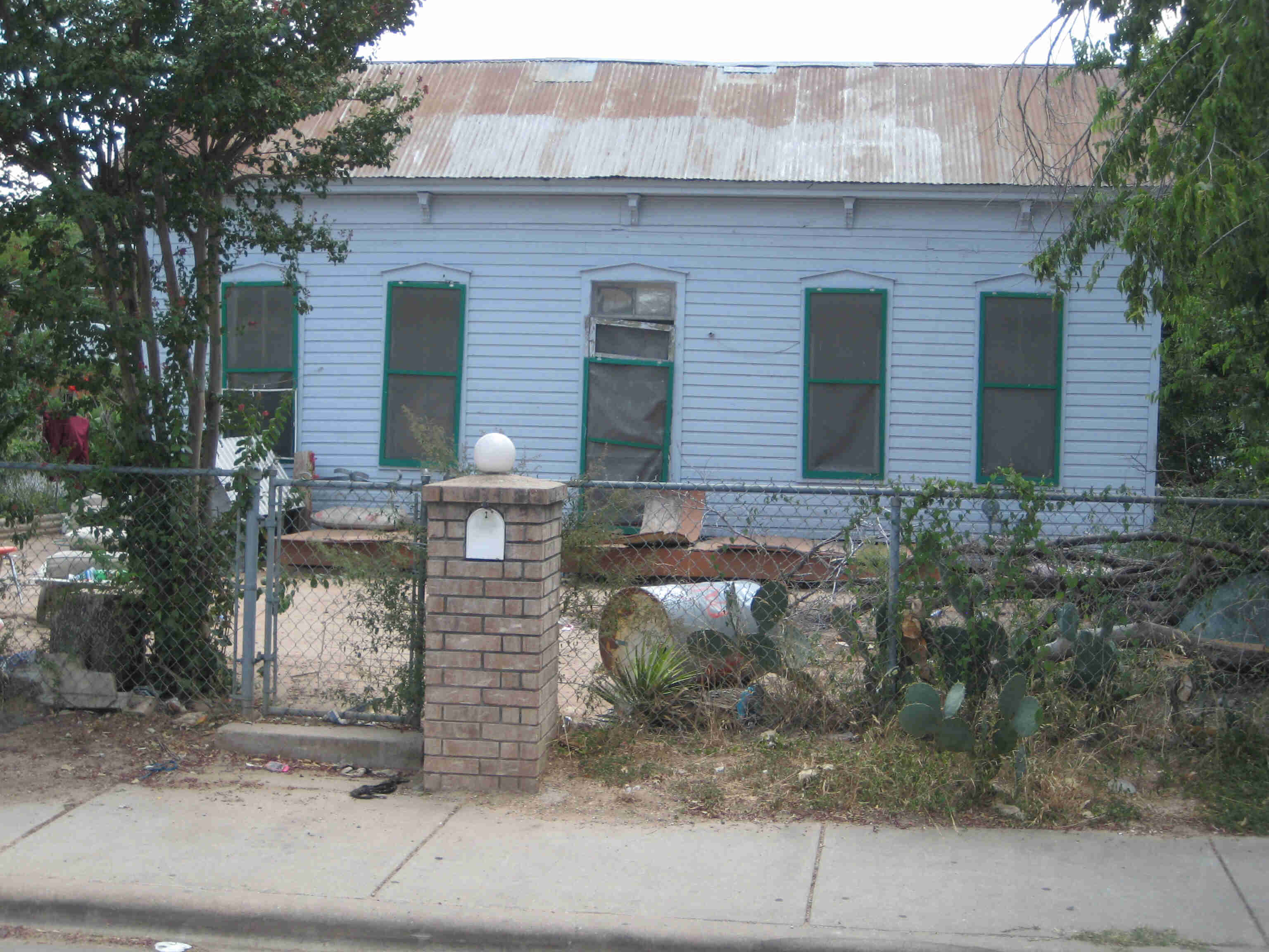 Front, street view of a house with a rusty steel roof and chain link fence around it, with junk laying around the yard