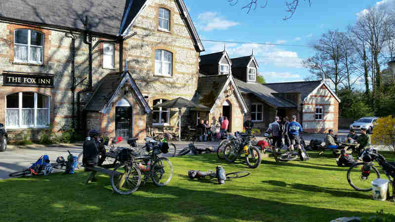 A group of cyclists with their bikes scattered around, in front of The Fox Inn building 