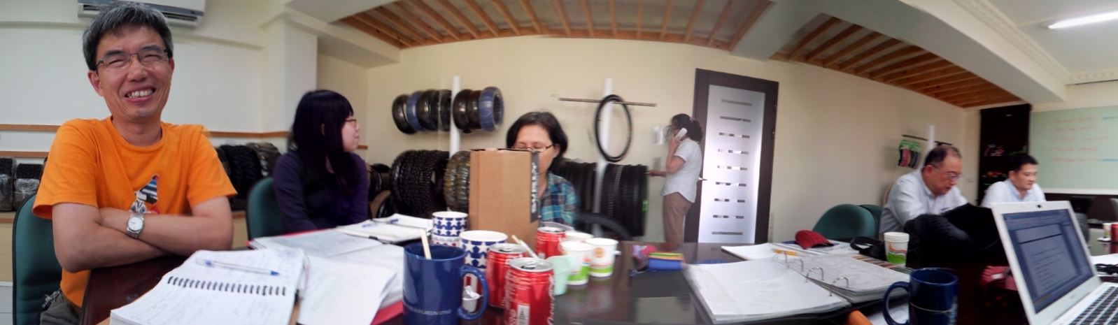 Three people sitting around a table, with notepads and beverages on it, in an office with bike tires in the background