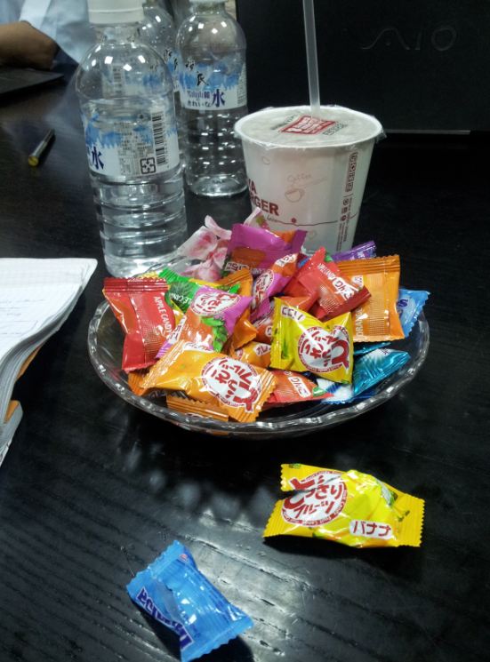 A glass tray with packages of candy in it, on a wood table, with a cup and bottles of water behind it