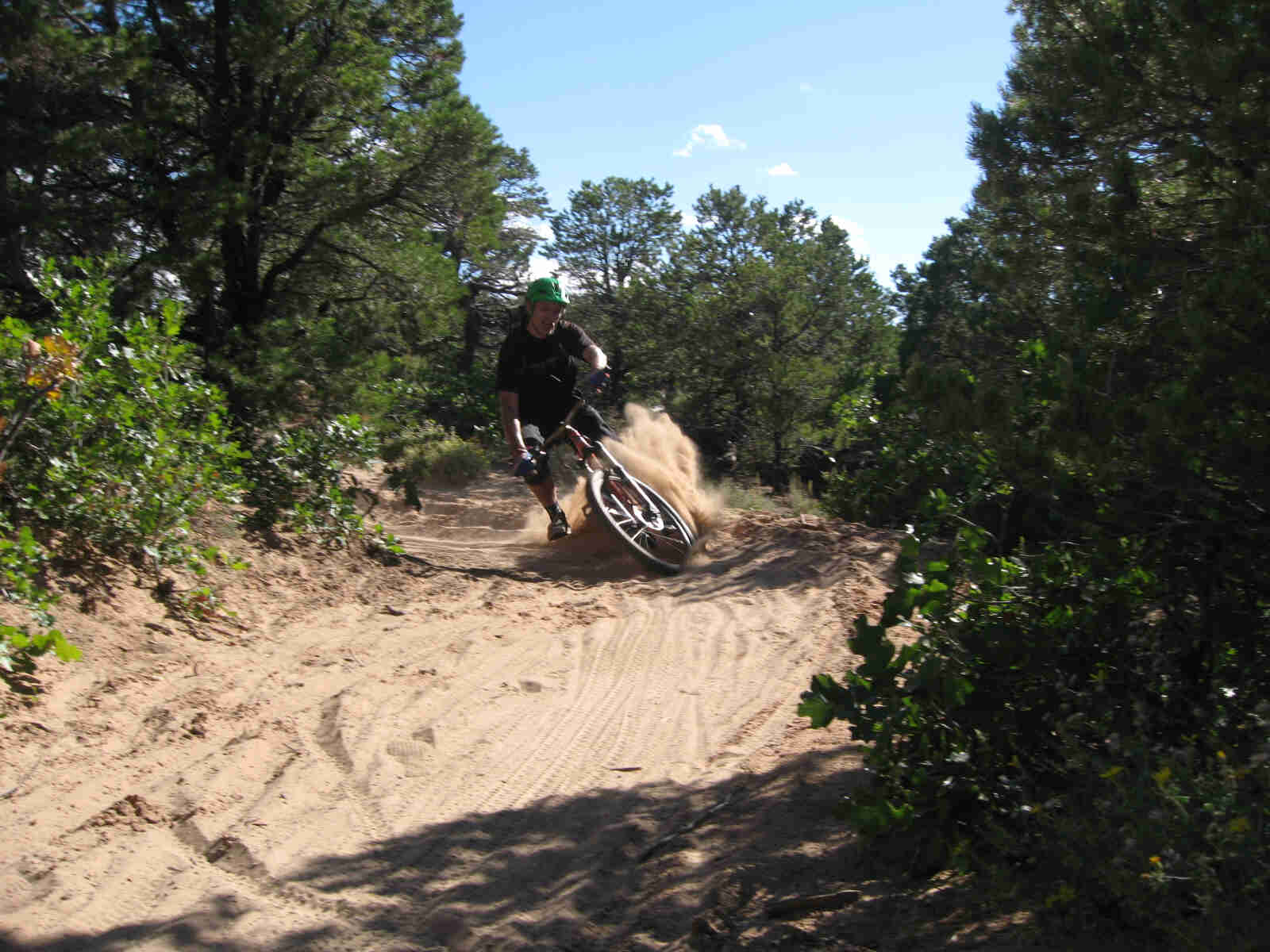 Front view of a cyclist, splashing up sand while rounding a corner on their Surly bike, on a trail with trees around it