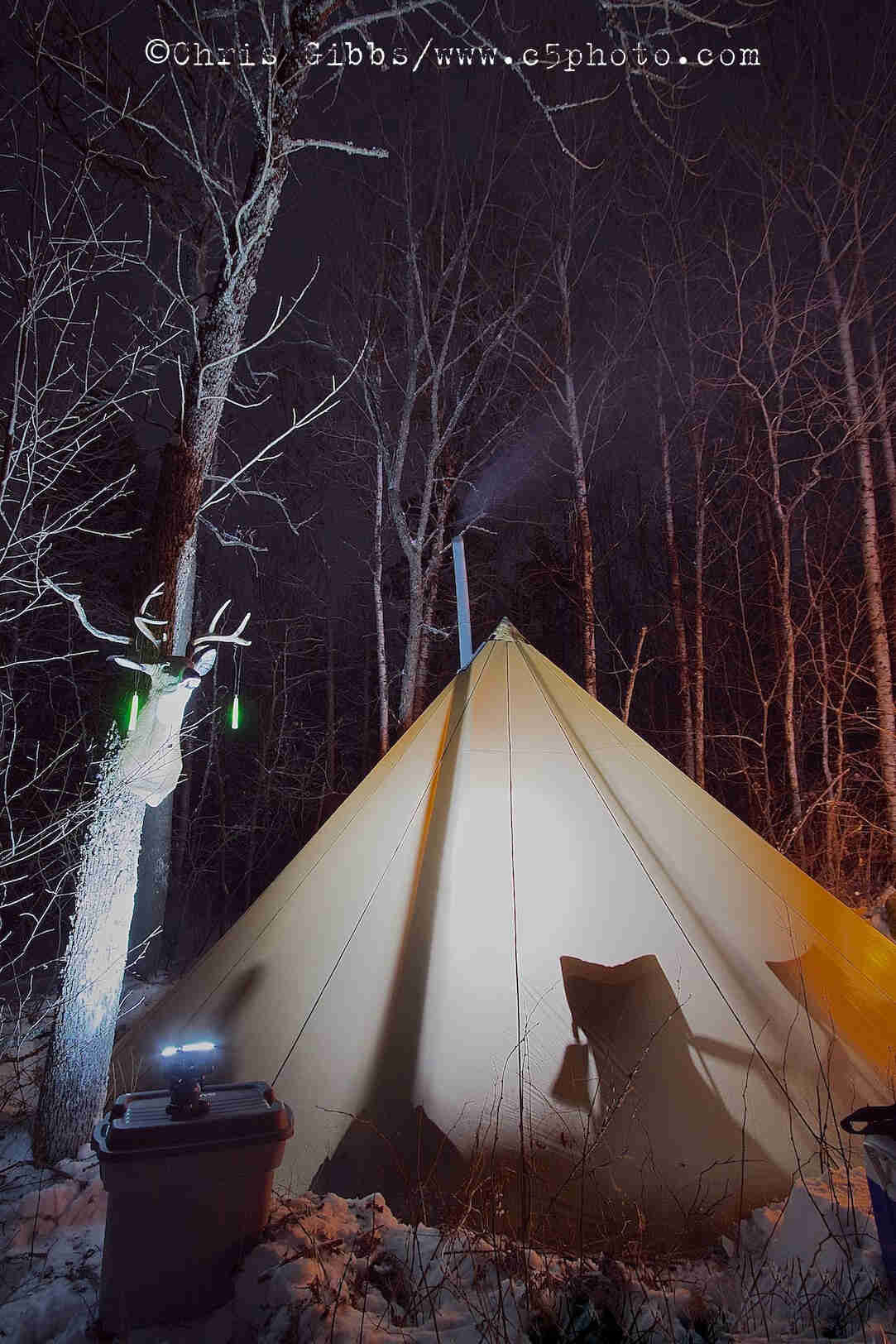 A teepee tent, lit up from the inside, on a snowy campsite in the woods at night