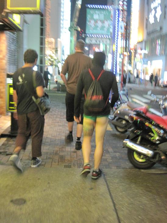 Rear view of 3 people, walking down a city sidewalk at night, with lit up buildings in the background