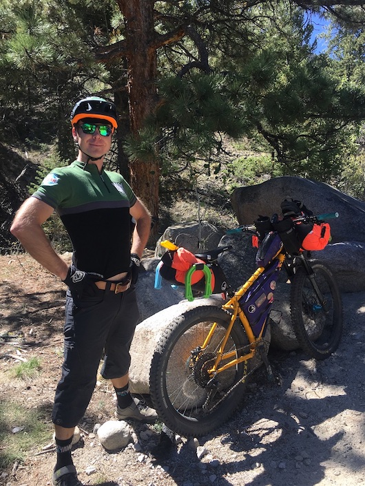 Cyclist stands at the back of a yellow Surly bike with gear leaning on a large rock, and pine tree in the background