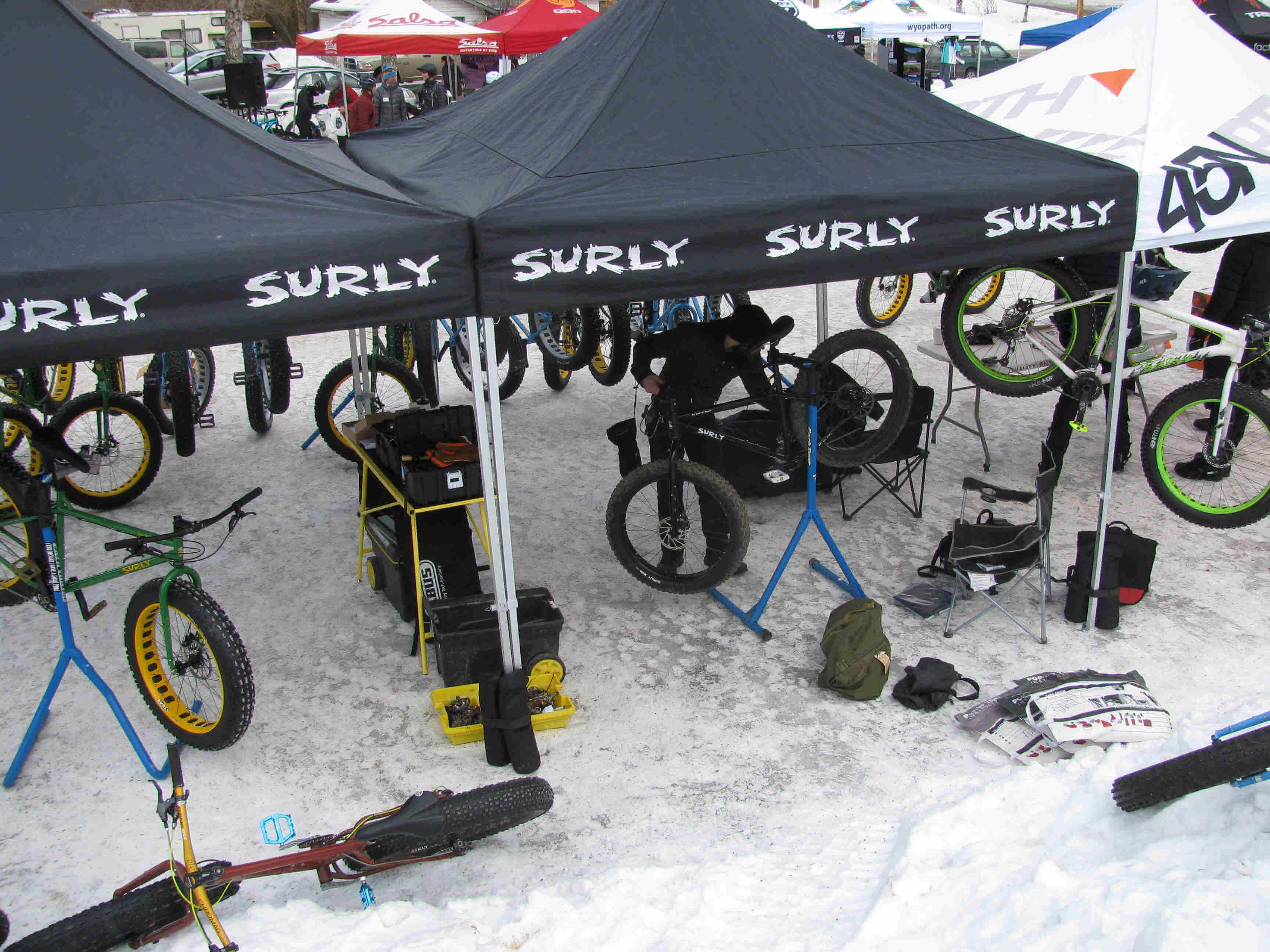 Downward view of a person, under a Surly Bikes canopy on snow, fixing a fat bike on a repair stand
