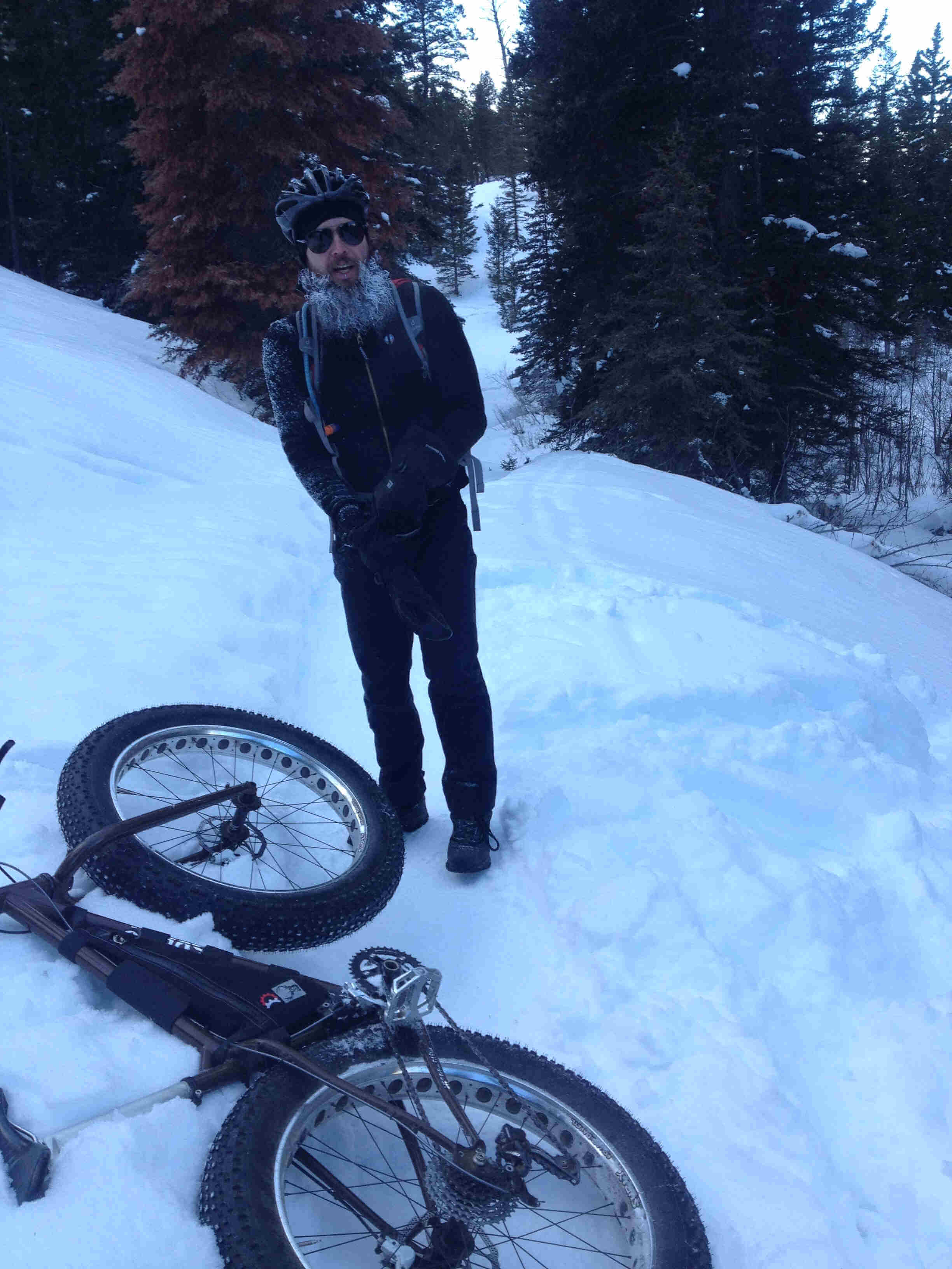 Front view of a cyclist with frozen beard, standing next to a Surly fat bike on it's side, on a snowy trail in a forest