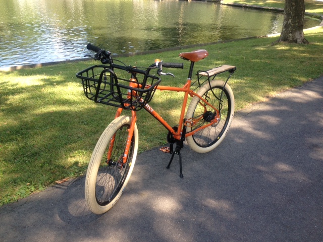 Left side view of an orange Surly Troll bike, parked on a paved trail, next to a grassy area with a pond behind it