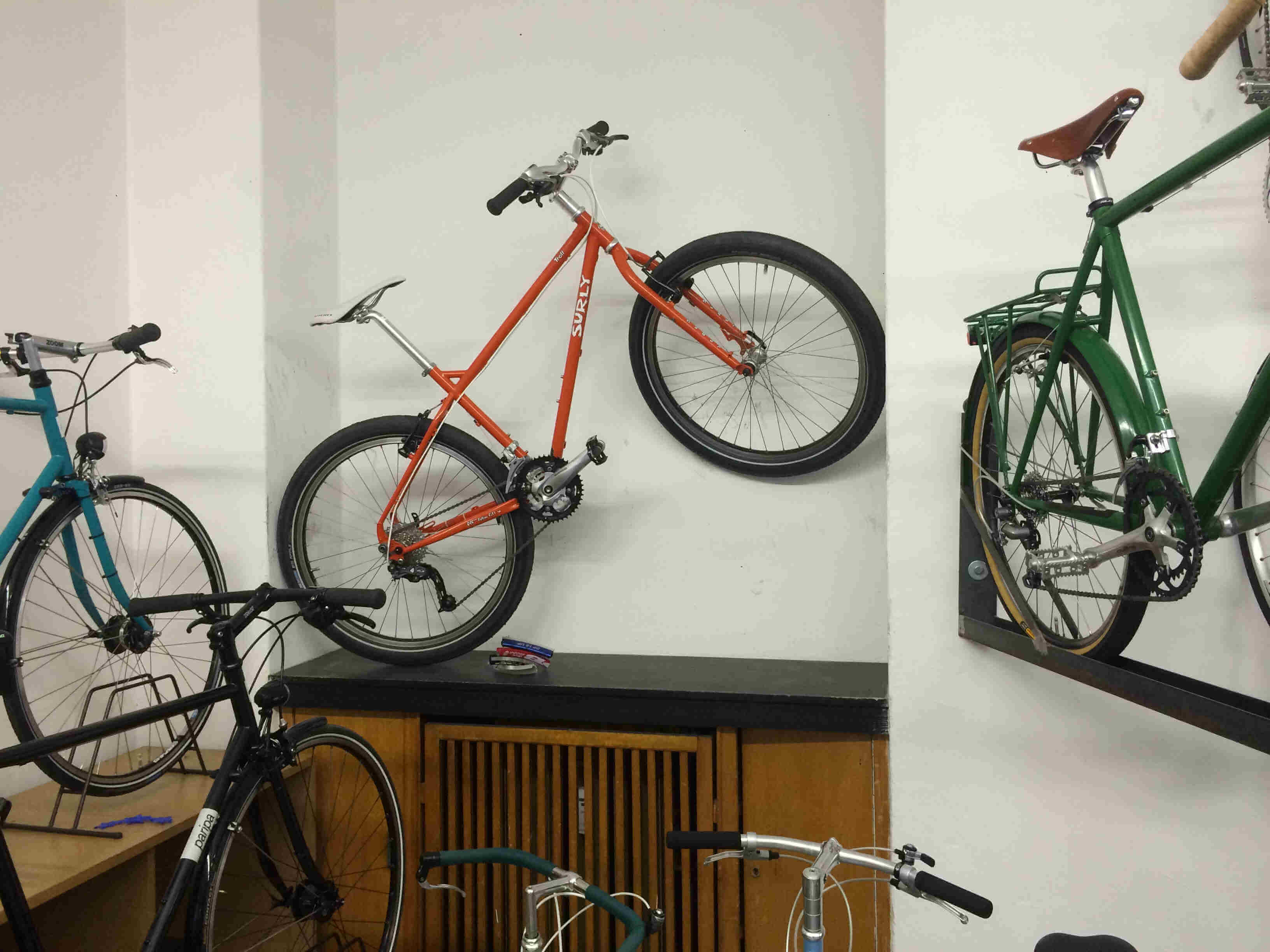 Right side view of an orange Surly Troll bike, wedged between 2 walls with front wheel propped up, in a bike shop room