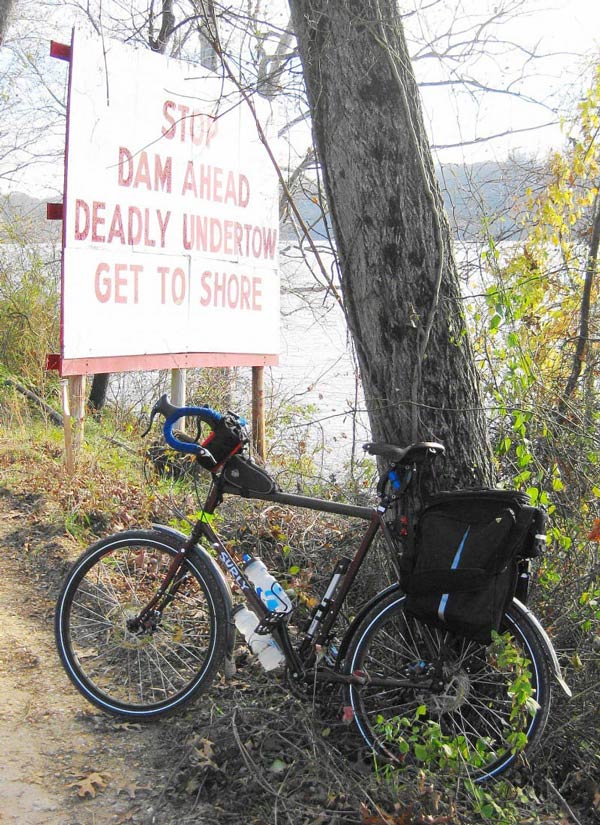 Left side view of a Surly Disc Trucker bike, on the side of a gravel road, with a sign and a river in the background