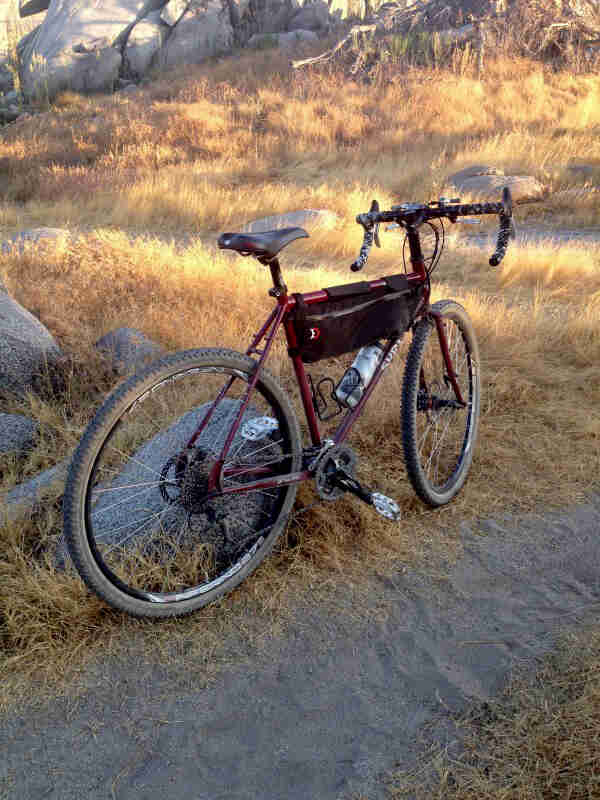 Rear, right side view of a red Surly Long Haul Trucker, parked along a sand trail in a brown grass field with rocks