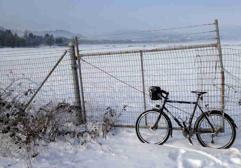 Left side view of a Surly Long Haul Trucker bike, leaning on a gate in a snow-covered field
