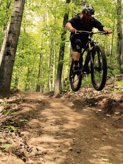Front view of a cyclist, riding a Surly Instigator bike, going airborne off a small jump on a dirt trail in the woods