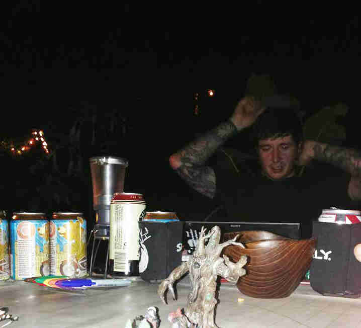 Front view of a person sitting behind a table with beer cans on it, looking down at a laptop computer at night