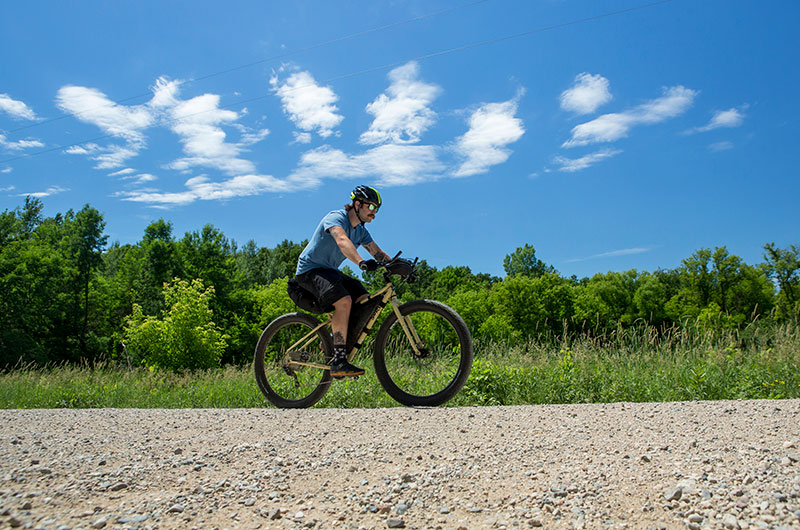 Person on sunny summer day riding loaded mountain bike with multi-position handlebar, trees in background