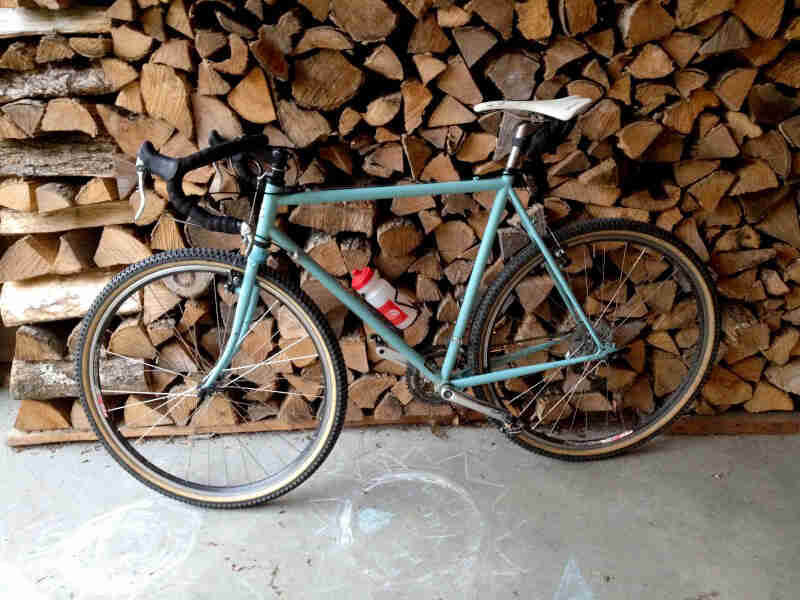 Left side view of a Surly Cross Check bike, mint, parked against a pile of split wood
