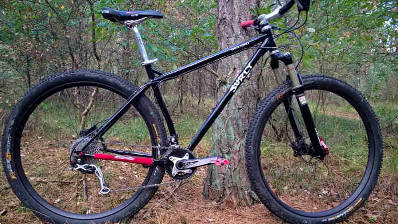 Right profile of a Surly Karate Monkey bike, black, parked on pine needles against a tree, with woods in the background
