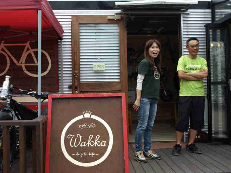 Front view of 2 people standing in front of an open door, outside of the Wakka bicycle life shop