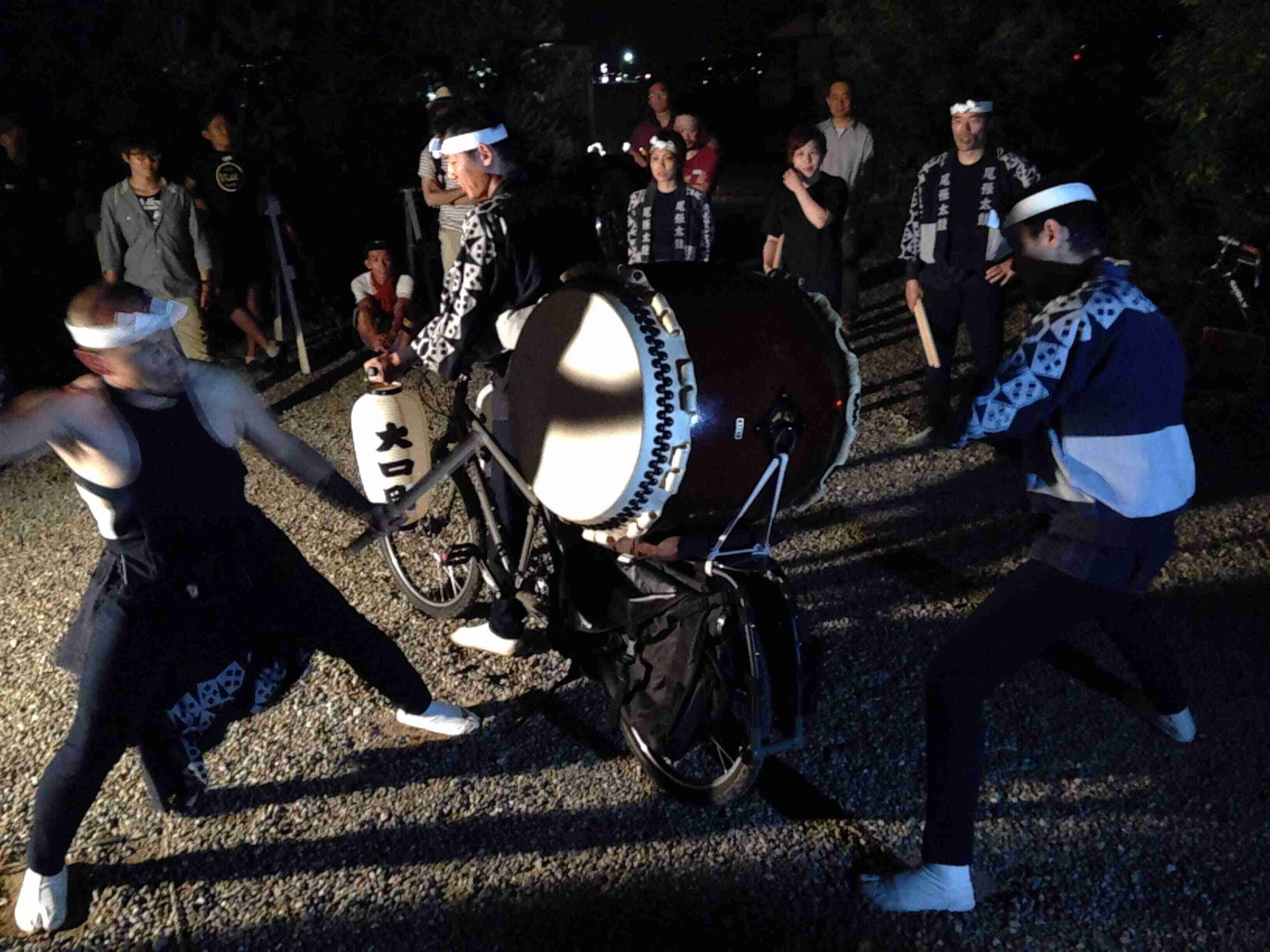 Two people beating on a large drum that's mounted horizontally on the back of a Surly Big Dummy bike, at nighttime