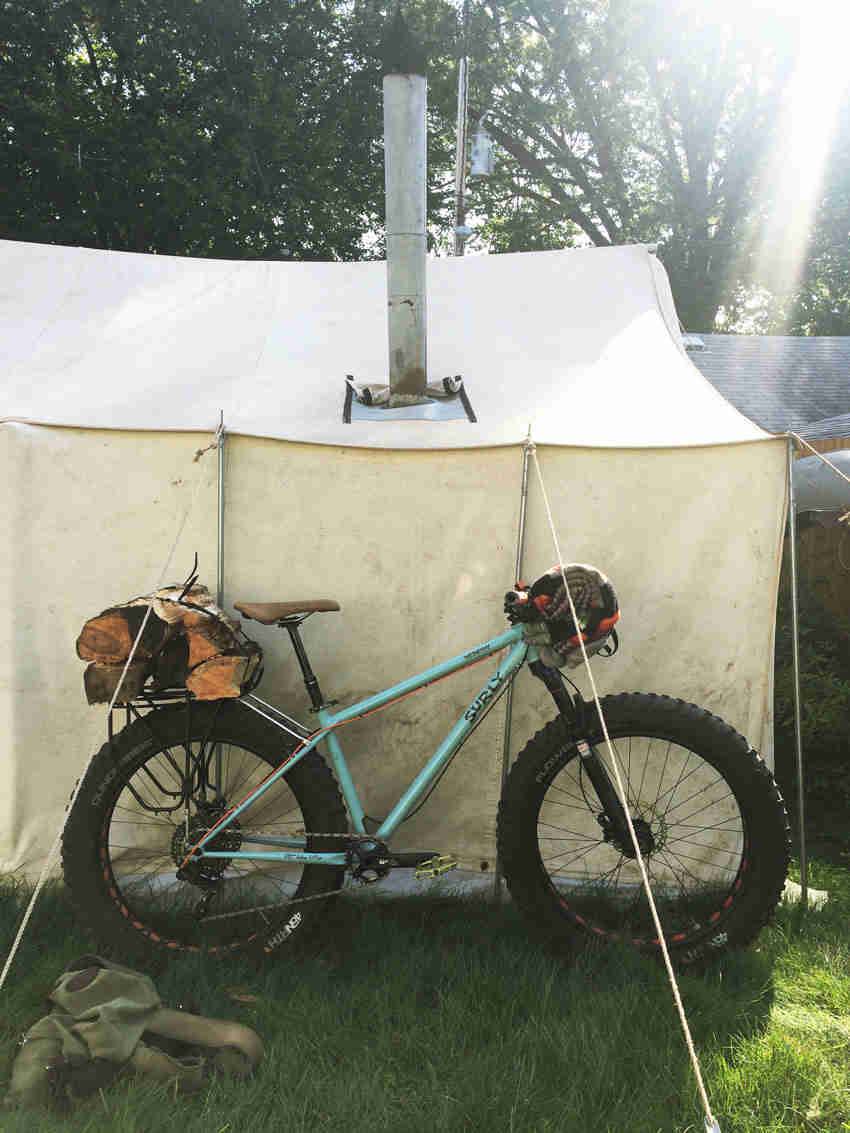 Right side view of a mint Surly Wednesday fat bike, with firewood on back, parked along a white tent with steel chimney