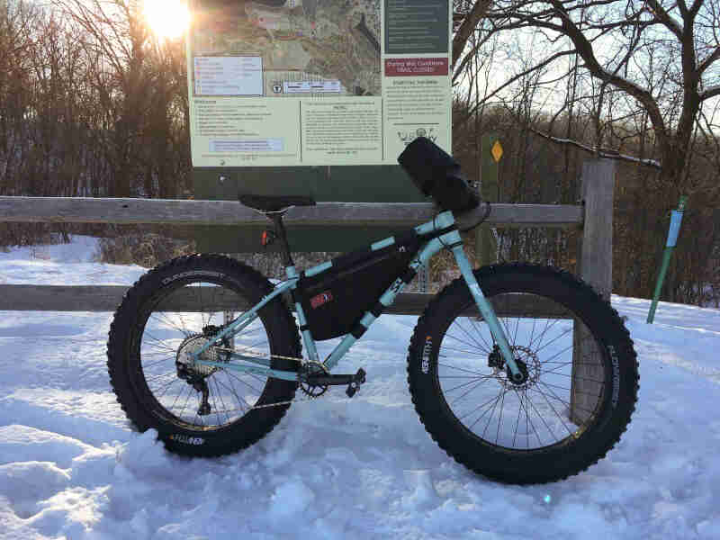 Right side view of a mint Surly Wednesday fat bike with gear packs, parked in the snow against a wood rail fence